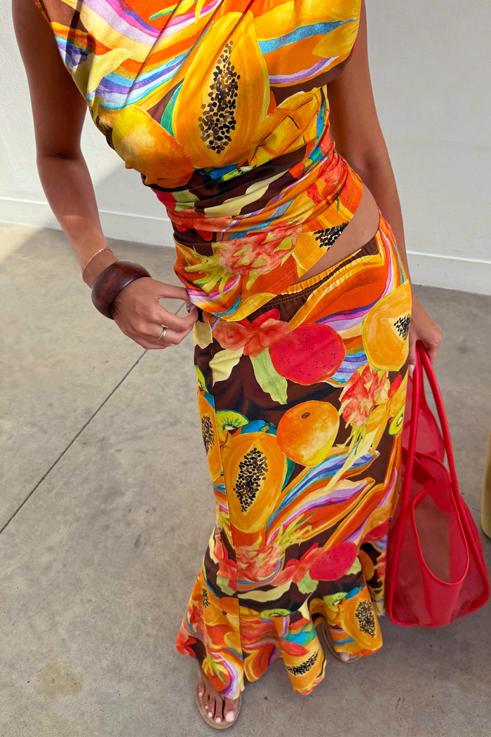 Rio A-Symmetric Skirt in a colourful tropical fruit print, styled with a matching top and red bag for a vacation look.