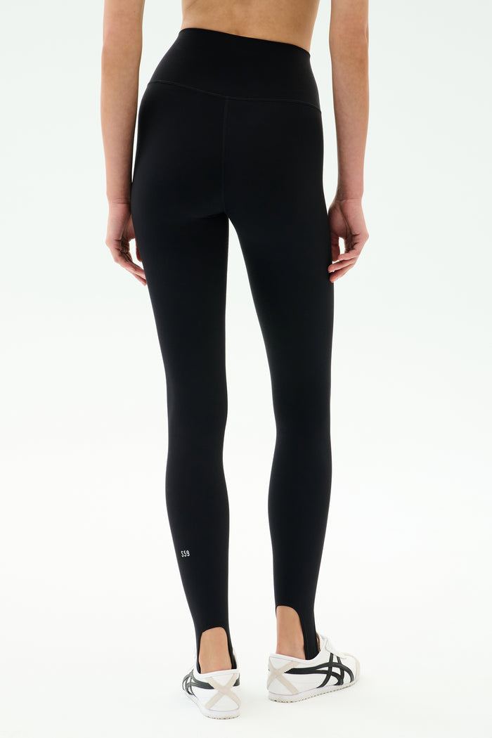 A person wearing SPLITS59 River High Waist Airweight Stirrup leggings in black with white athletic shoes, shown from the back against a plain background.