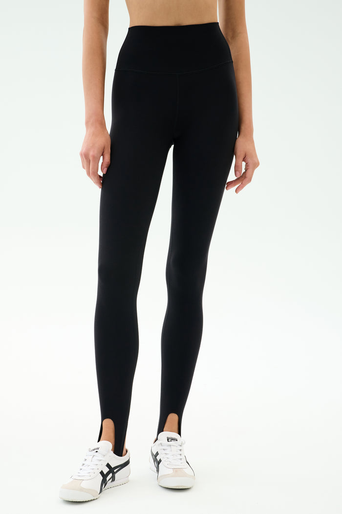 A person wearing SPLITS59 River High Waist Airweight Stirrup leggings in black with white sneakers, standing against a plain light background.