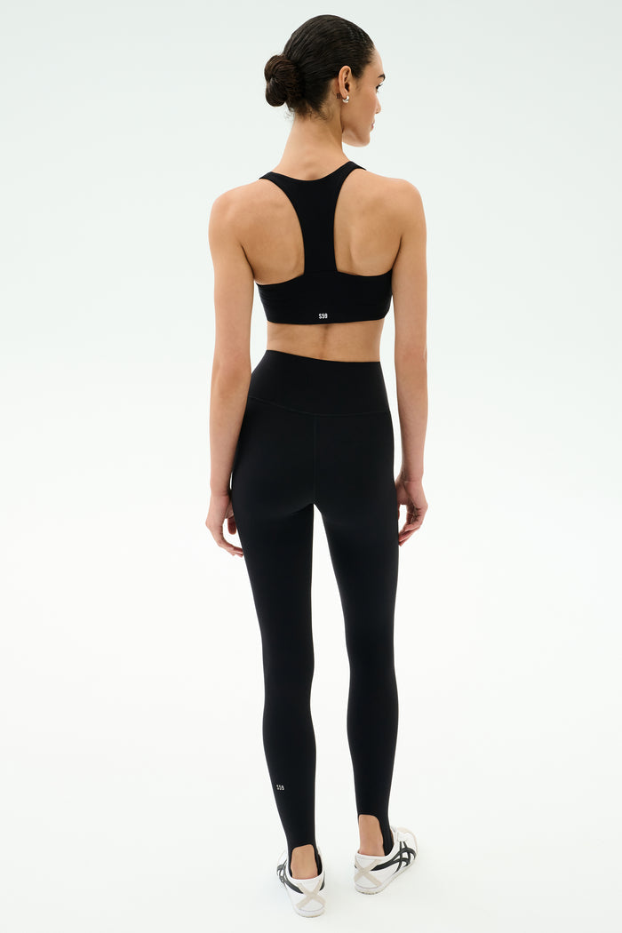 A woman faces away, wearing SPLITS59’s River High Waist Airweight Stirrup leggings in black with a matching sports bra and white sneakers, set against a plain white background.