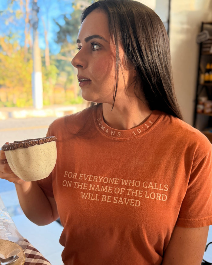 A woman wearing an orange crewneck t-shirt with a white printed Bible verse, holding a small ceramic bowl in a brightly lit indoor setting.