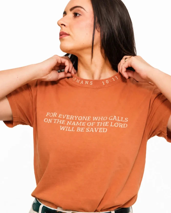 A woman wearing a rust-orange crewneck t-shirt with a white printed Bible verse from John 10:13 on the chest, posing against a white background.