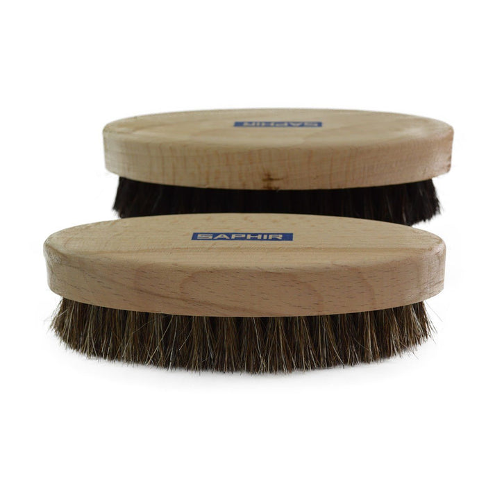 Two oval shoe shine brushes with horsehair bristles, one with black bristles and one with natural brown bristles, both with wooden handles, on a white background.