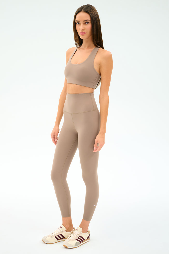 A woman in a SPLITS59 Sara Airweight Bra in Latte stands against a plain white background, ready for Pilates or hot yoga.