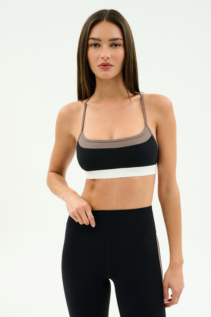 A woman stands against a plain background wearing the SPLITS59 Scottie Airweight Bra in Black/Lentil with white trim and matching high-waisted leggings, both crafted from soft nylon and spandex for a comfortable, flexible fit.