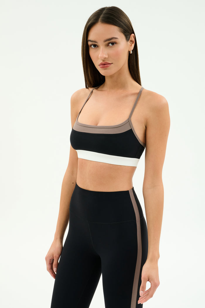 A woman in SPLITS59's Scottie Airweight Bra—Black/Lentil—made from a nylon-spandex blend, with brown and white accents, wears matching high-waisted leggings against a plain light background.