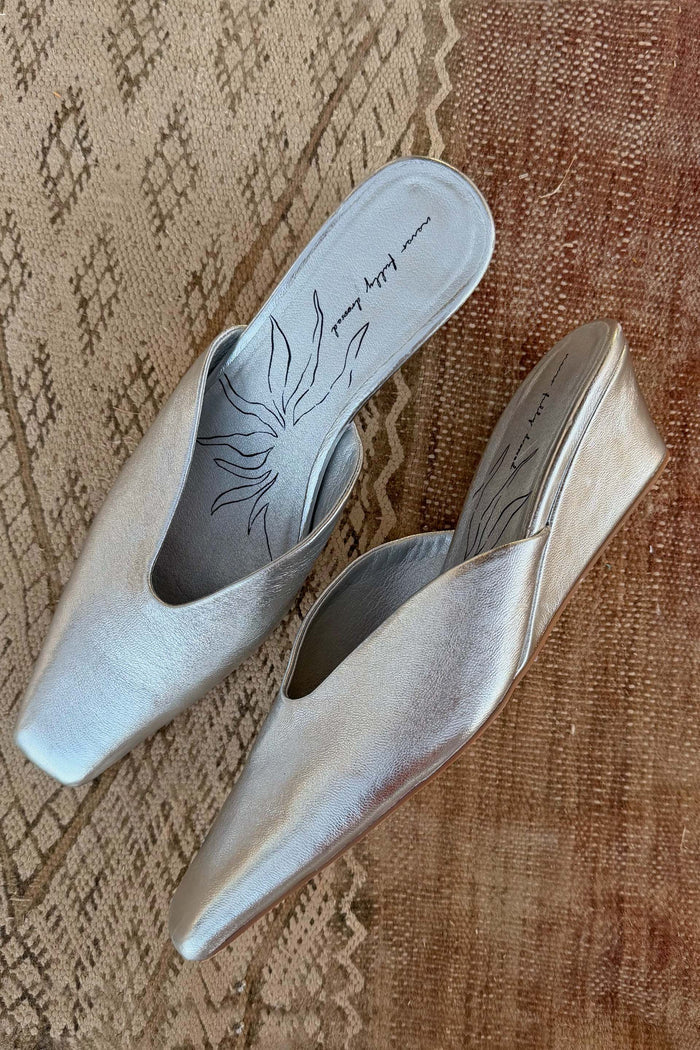 Silver metallic leather wedge mules featuring a pointed toe and sun-printed insole, resting on a patterned rug.