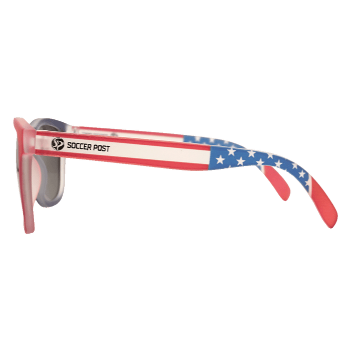 A side view of sunglasses with an American flag design on the temples. The temples feature red and white stripes with a blue, star-patterned ear piece, and the "Soccer Post" logo and name printed on the side. The front frame is red with dark lenses.