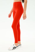 A person wearing SPLITS59 Sprint High Waist Rigor 7/8 leggings in Lotus Red and white athletic shoes with black stripes, standing against a plain light background.