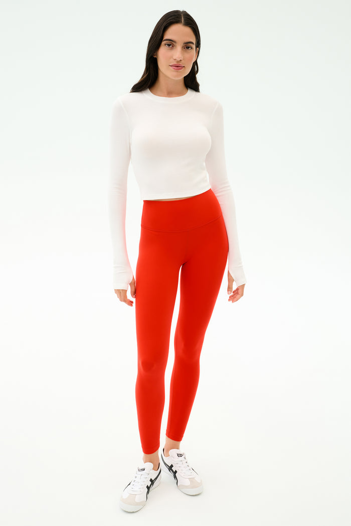A person stands facing forward in a white long-sleeve top, white sneakers, and SPLITS59 Sprint High Waist Rigor 7/8 leggings in Lotus Red, against a plain white background.