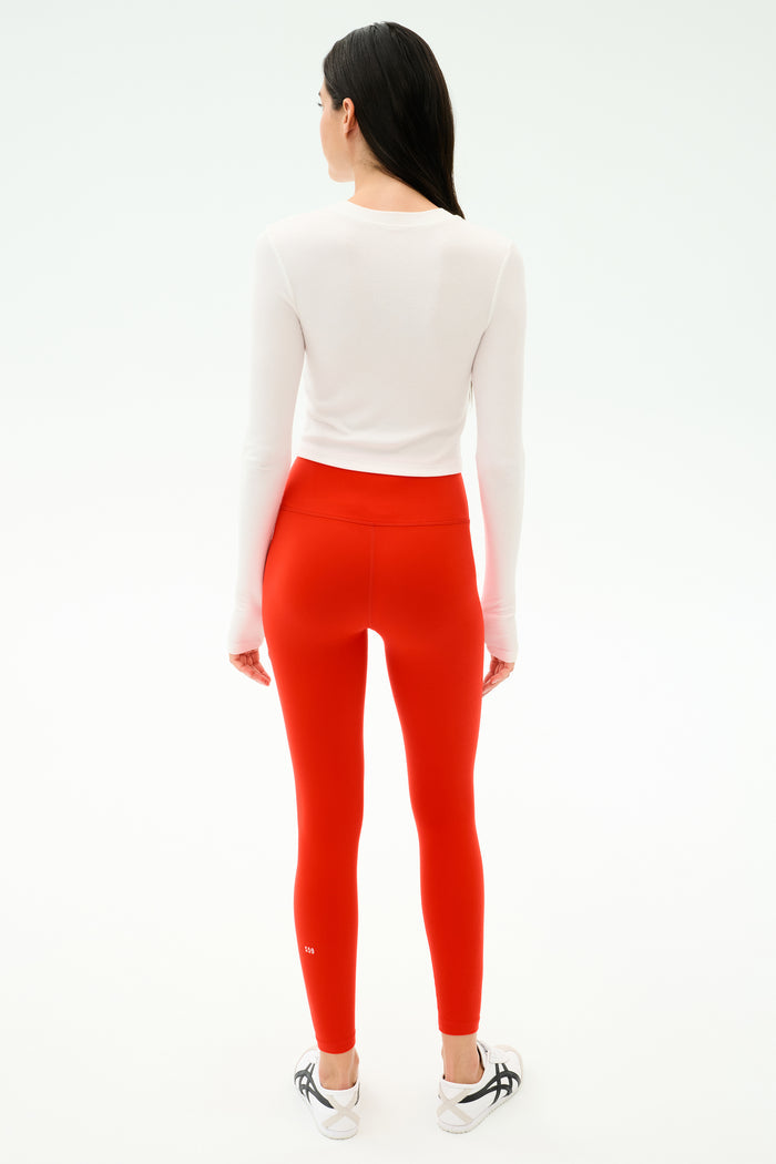 A woman faces away, wearing SPLITS59 Sprint High Waist Rigor 7/8 leggings in Lotus Red and a white long-sleeve top, paired with white sneakers with black accents, all set against a plain white background.