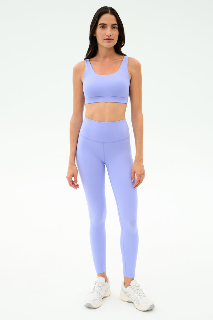 A woman stands facing forward in the SPLITS59 Sprint Rigor Bra - Purple Haze and matching leggings with white athletic shoes, designed for multi-sport performance, against a plain white background.