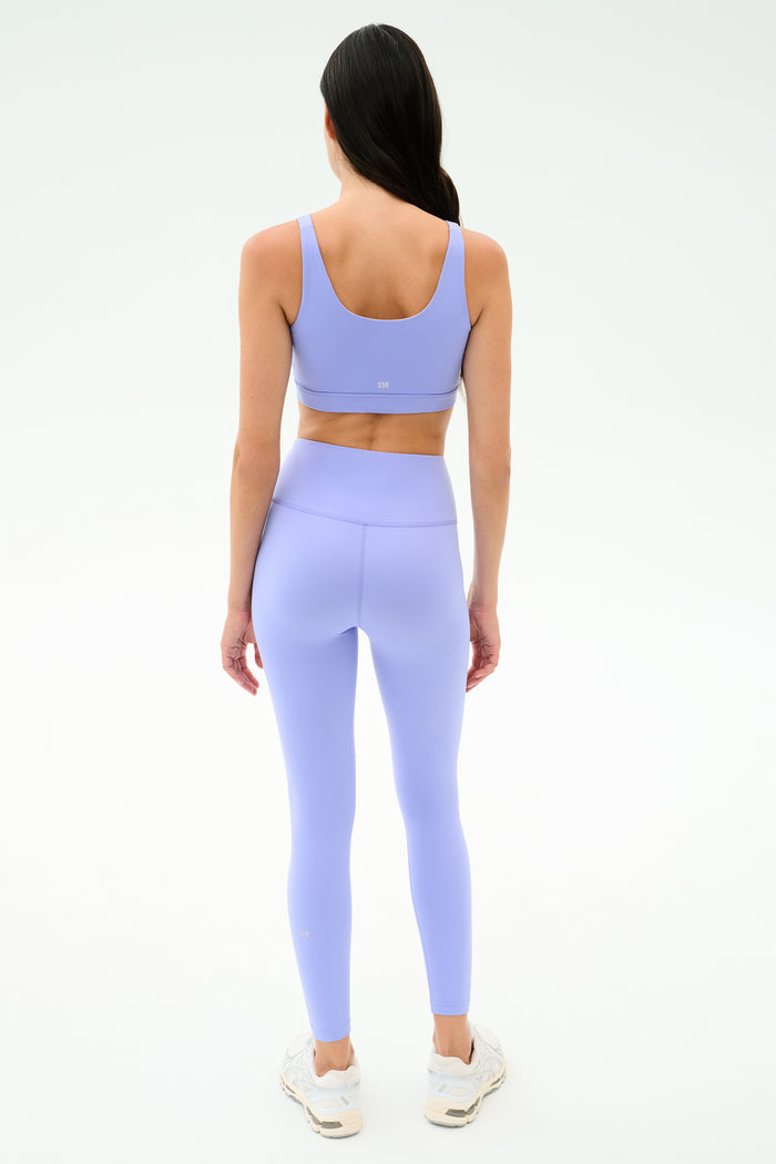A woman, seen from behind, wears SPLITS59 Sprint High Waist Rigor 7/8 leggings in Purple Haze with a matching sports bra and white sneakers against a plain light background.