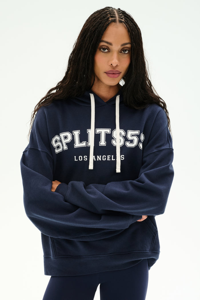 A woman stands with arms crossed against a plain background, wearing the SPLITS59 Stella French Terry Oversized Hoodie in Indigo—a soft 100% cotton piece featuring "SPLITS59 LOS ANGELES" printed on the front.