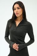 A woman with long dark hair wears the SPLITS59 Stevie Active Rib Half Zip Waist Length top in Heather Graphite, standing against a plain light background.