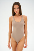 A woman wearing the Suki Airweight Bodysuit in Latte by SPLITS59 stands against a plain background, facing forward with a neutral expression.