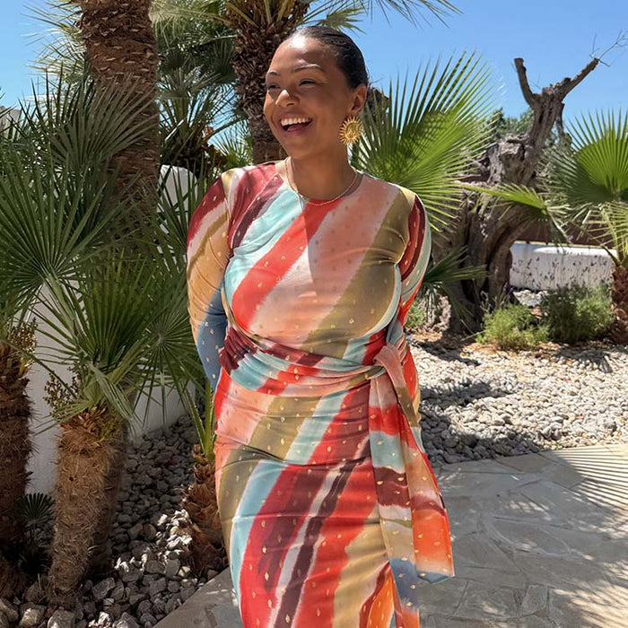 Sunset Stripe Mesh Midi Dress with a multicoloured stripe print and tie waist, worn in a sunny palm tree setting.