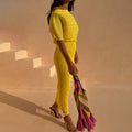 Yellow plissé Sunshine Tilly Dress with gold flecks, styled off-the-shoulder with a colourful tassel bag.