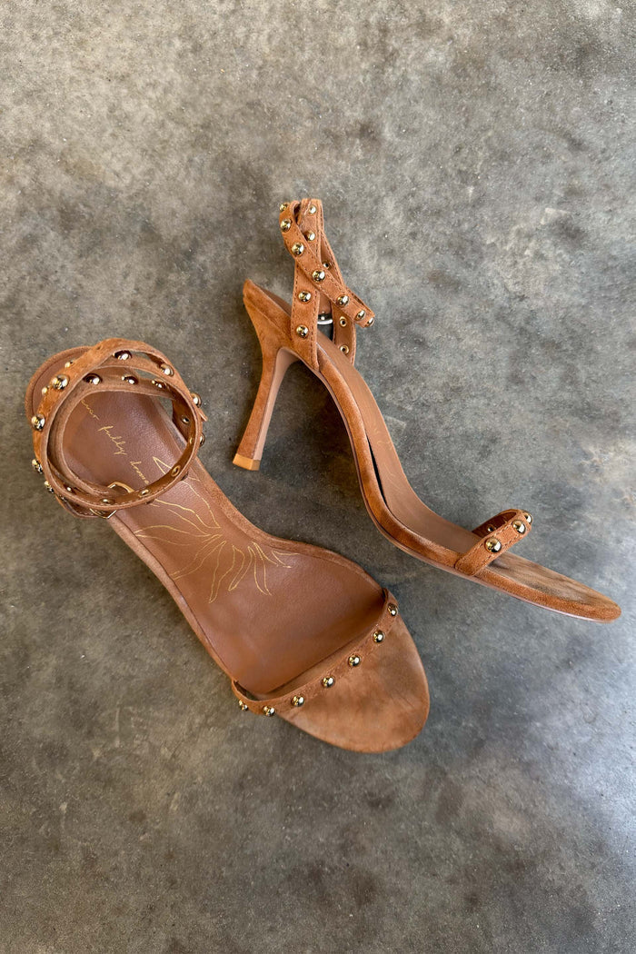 Tan suede stiletto heels with gold stud detailing and wrap-around ankle straps shown on a grey textured floor.