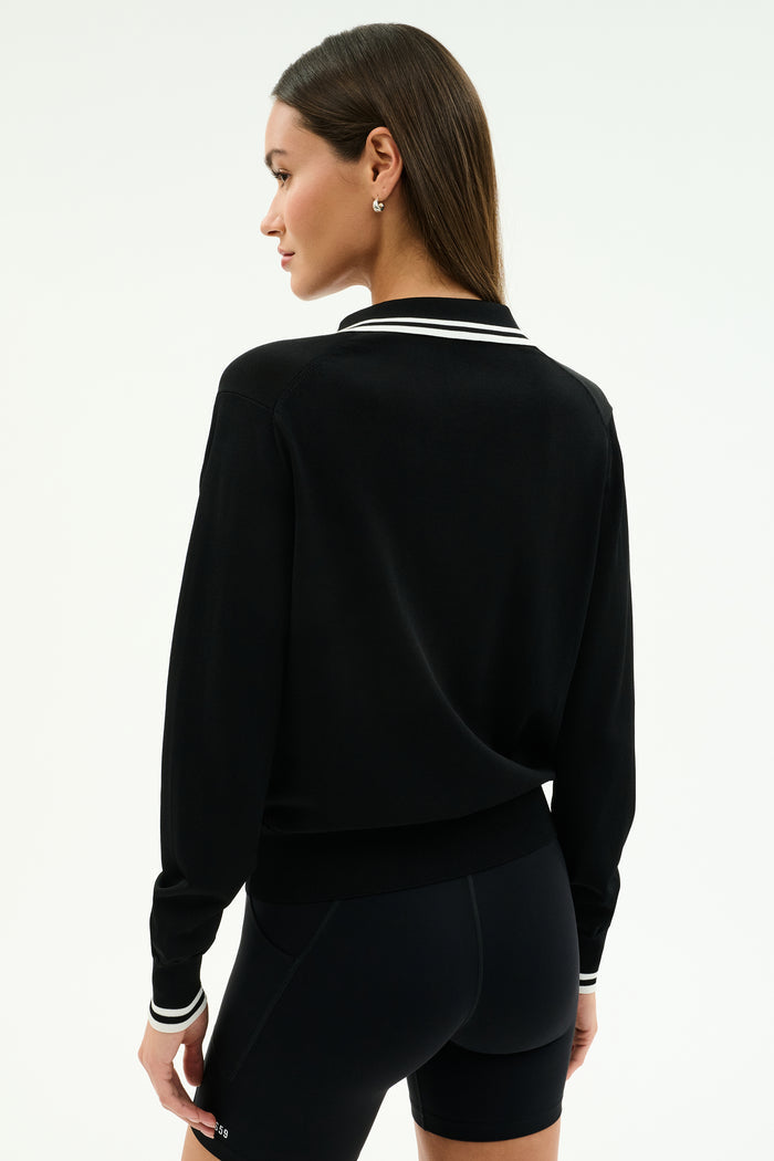 A woman stands with her back to the camera in the SPLITS59 Taylor Knit Polo in Black/White, a soft rayon-nylon blend long-sleeve top featuring white trim, styled with black fitted shorts against a plain light background.