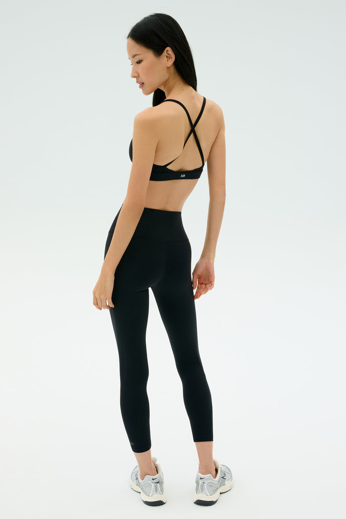 A woman wears the SPLITS59 Theo Rigor Bra - Black with black leggings and gray running shoes, standing and looking over her shoulder. Ideal for multi-sport performance, set against a plain background.