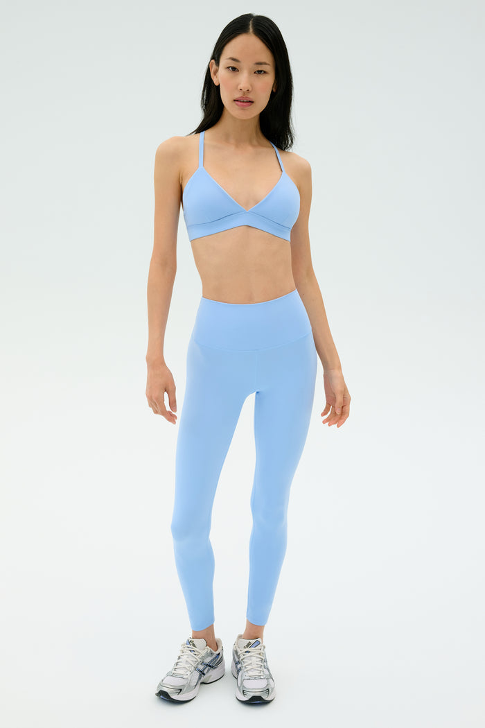 A woman faces forward wearing the SPLITS59 Theo Rigor Bra in Skylight with matching leggings and silver athletic shoes, set against a plain light background.
