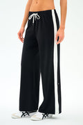 A person models the SPLITS59 Willow Airweight Wide Leg pants in Black/White, featuring buttery-soft fabric, wide legs, white side stripes, and a white drawstring, paired with white sneakers against a plain background.