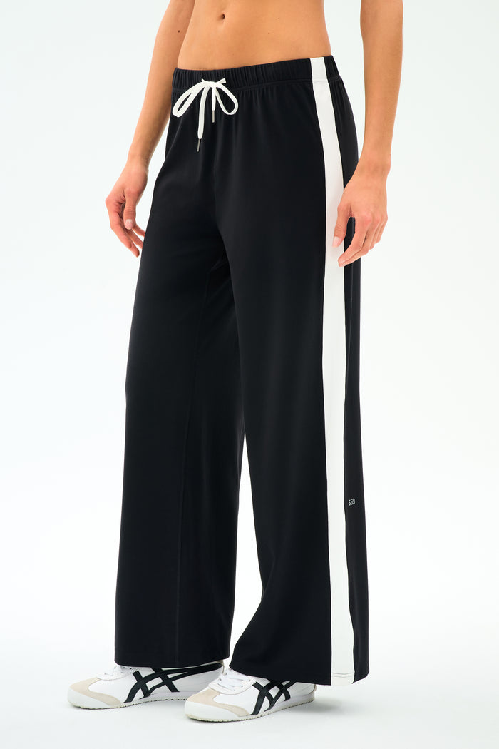 A person models the SPLITS59 Willow Airweight Wide Leg pants in Black/White, featuring buttery-soft fabric, wide legs, white side stripes, and a white drawstring, paired with white sneakers against a plain background.