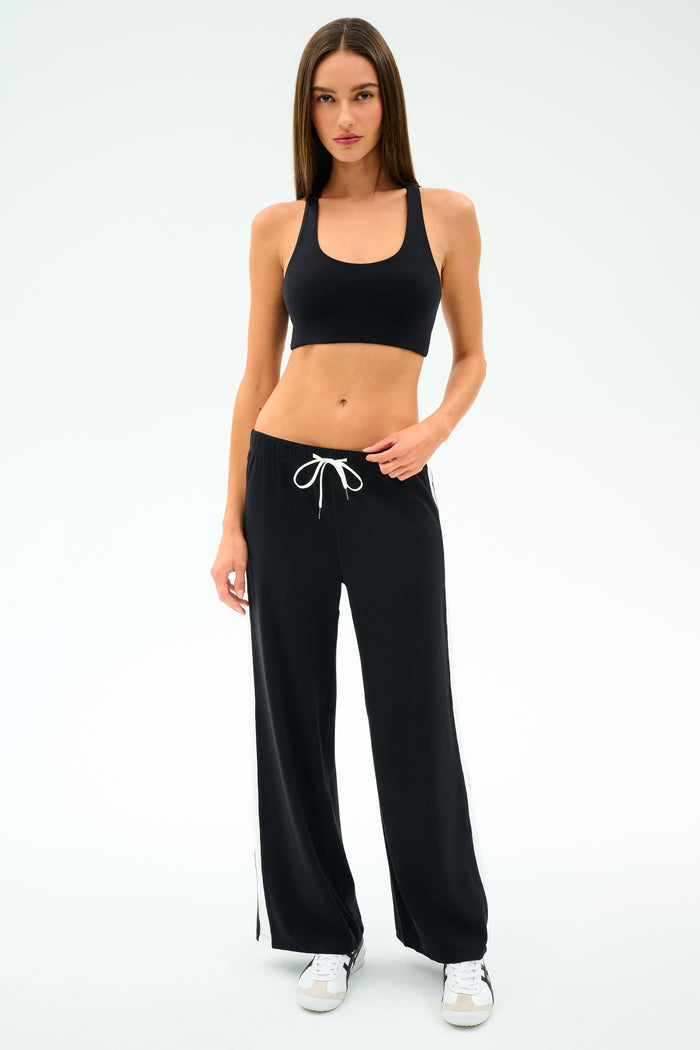 A woman wearing the SPLITS59 Willow Airweight Wide Leg pants in Black/White with a black sports bra and white sneakers stands against a white background.