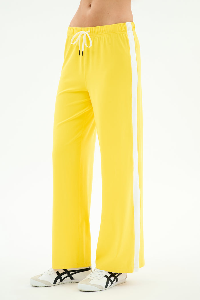 A person wears SPLITS59's Willow Airweight Wide Leg pants in bright yellow/white with white sneakers featuring black accents, standing against a white background.