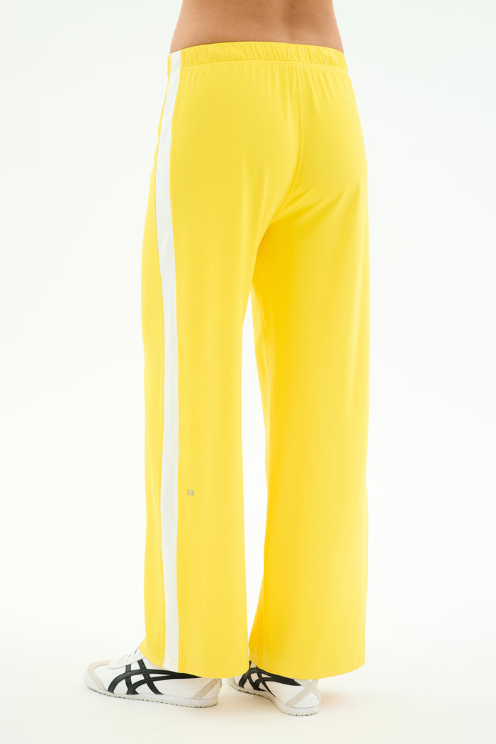 Person wears SPLITS59 Willow Airweight Wide Leg pants in Bright Yellow/White, featuring a white side stripe and soft spandex blend, paired with white sneakers, shown from the back against a plain background.