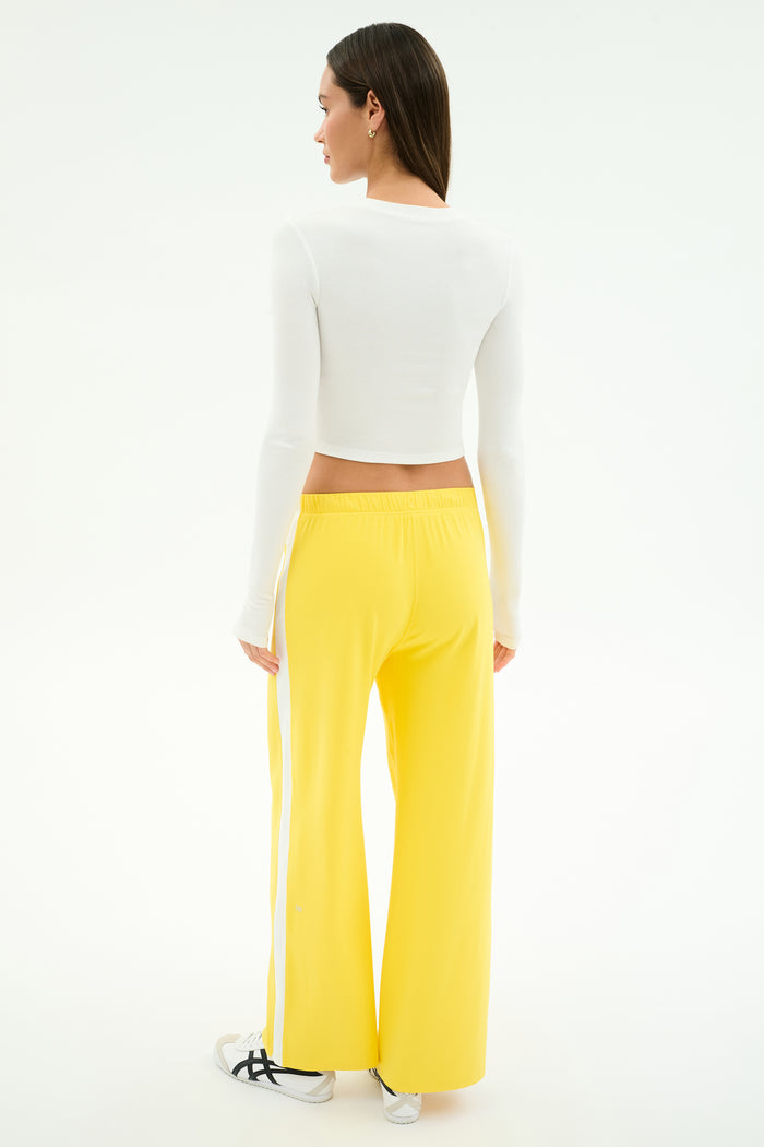 A person stands facing away, wearing SPLITS59's Willow Airweight Wide Leg pants in Bright Yellow/White and a long-sleeve white crop top, paired with white sneakers with black stripes, against a plain white background.